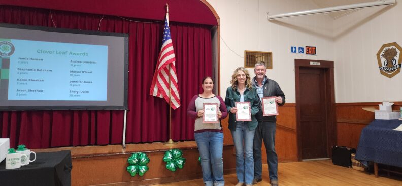 Three smiling adults stand side by side next to a small stage holding certificates during a 4-H awards event. Behind them is a red curtain and an American flag. To the left, a projector screen displays the “Clover Leaf Awards” list of names.
