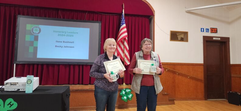 Two women stand side by side next to a small stage holding certificates during a 4-H awards event. Behind them is a red curtain and an American flag.