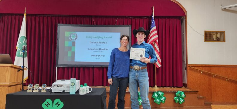 A smiling woman and a young person in a cowboy hat stand on a small stage in front of a red curtain. The young person is holding a certificate.