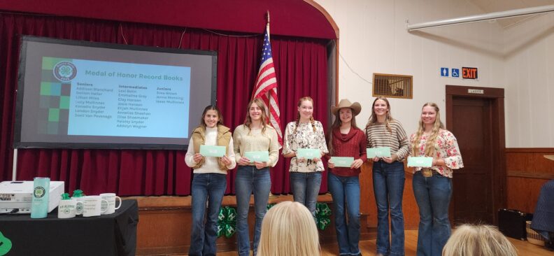 Six young girls smiling, standing side by side next to a small stage holding green envelopes during a 4-H awards event. Behind them is a red curtain and an American flag. To the left, a projector screen displays the “Medal of Honor Record Books” list of names.