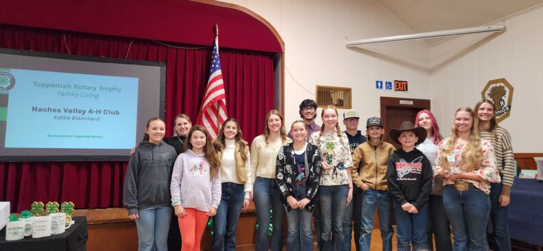 A group of kids proudly while standing side by side displaying their two club awards (trophies), during a 4-H awards event. Behind them is a red curtain and an American flag. To the left, a projector screen displays the “Toppenish Rotary Trophy” "Naches Valley 4-H Club"