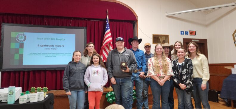 A group of kids proudly while standing side by side displaying their club award (trophy), during a 4-H awards event. Behind them is a red curtain and an American flag. To the left, a projector screen displays the “Inez Walters Trophy” "Sagebrush Riders"