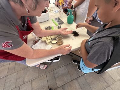 Young boy planting a seed in a clear CD case while being helped by Master Gardener Reggie at the Farmer's Market Kids' table