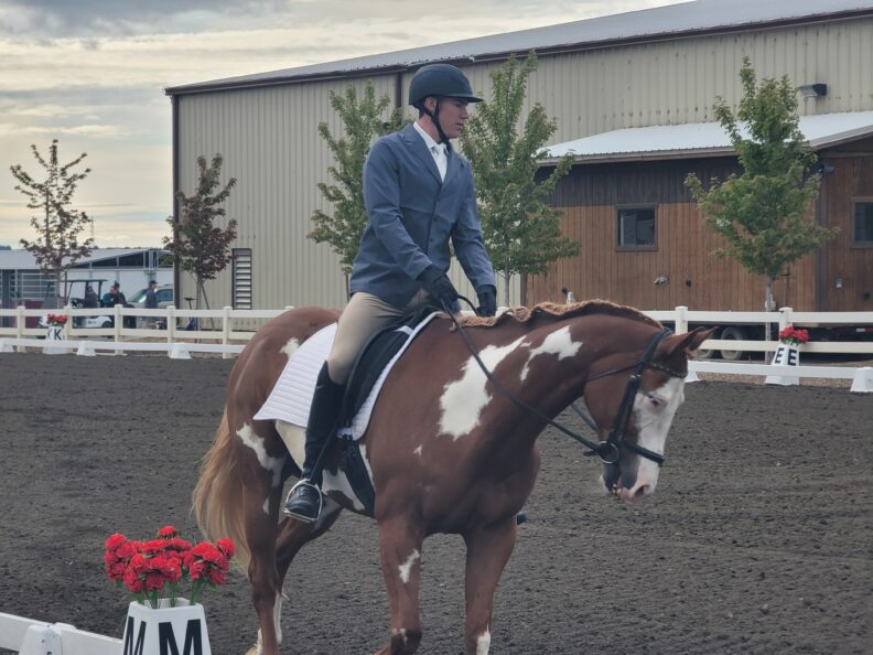 A male rider in show attire rides a paint horse in an outdoor dressage arena, passing a dressage marker and red flowers.