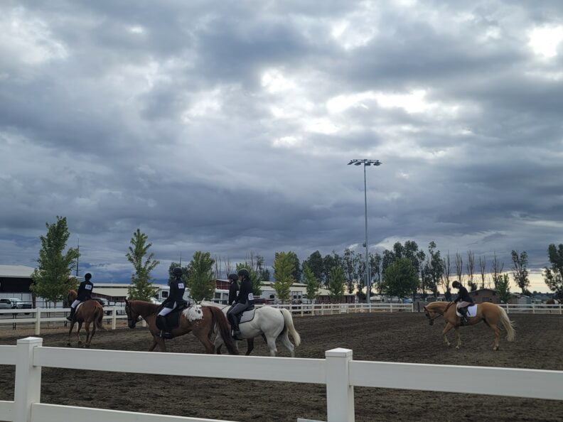 Several riders warm up their horses in an outdoor arena beneath a cloudy sky, before an equestrian event.