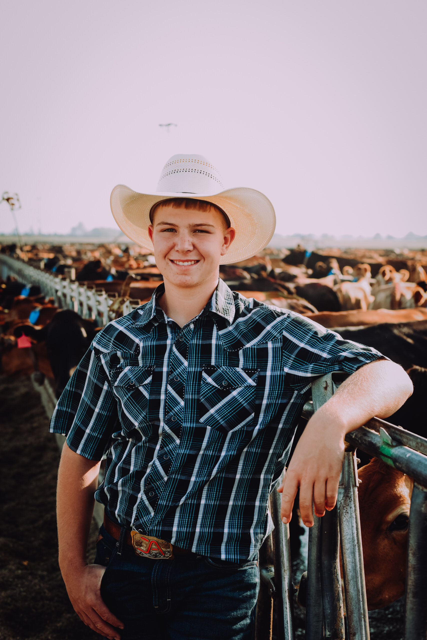 Teenage boy in a blue patterned button up shirt and cowboy hat, smiling while posing along side the metal gate of a dairy farm.