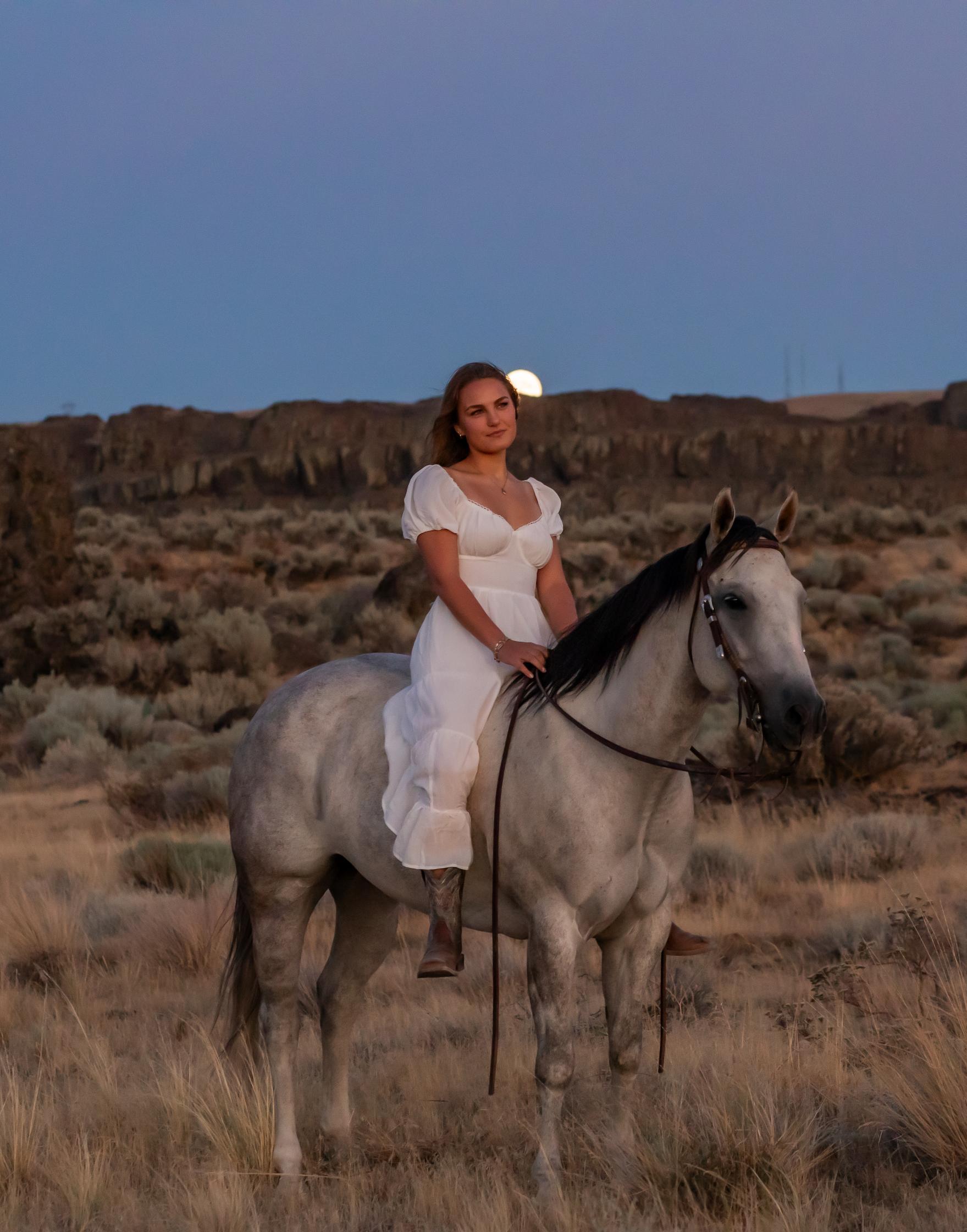 A teenage girl wearing a white dress sits on a gray horse with a black mane in a sagebrush field. She is looking into the distance, with mountains visible in the background.
