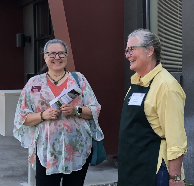 Two Master Gardeners having a conversation while smiling.  One holds a flier, the other wears a dark green work apron.