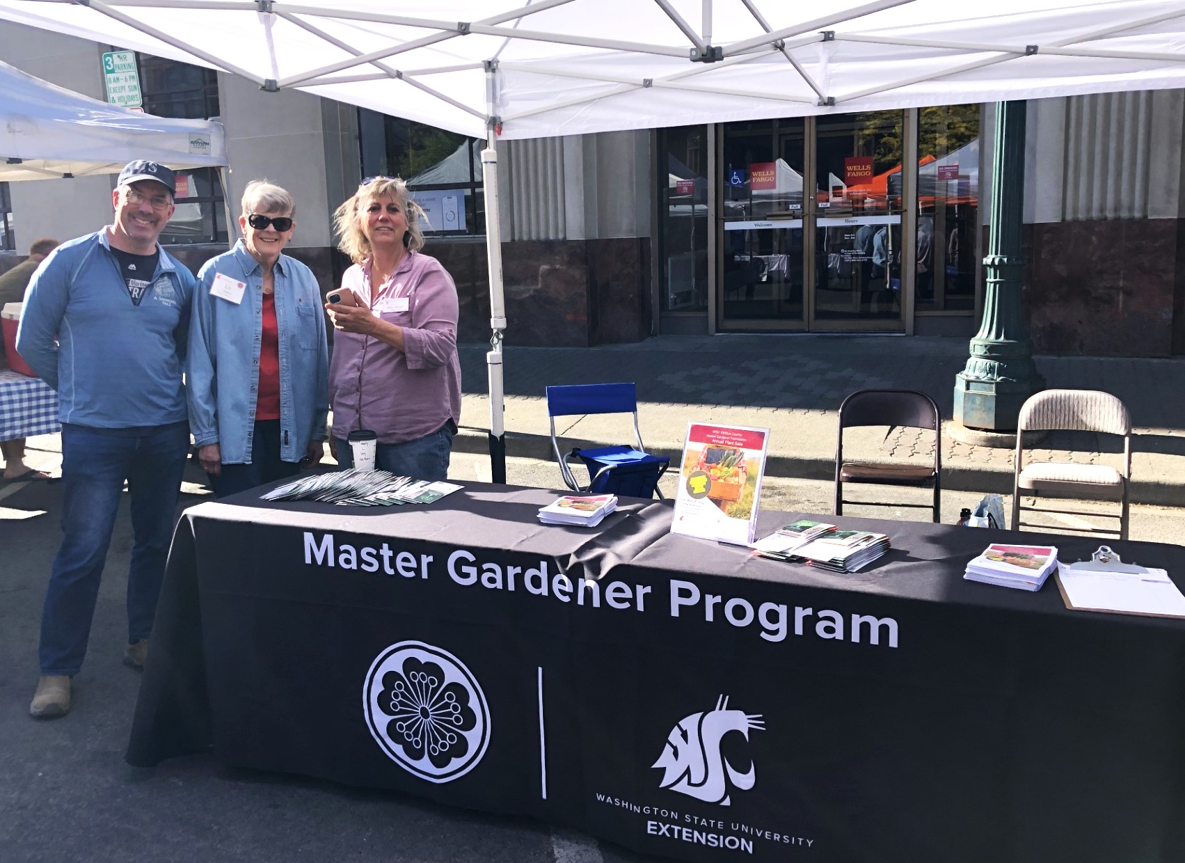 Master Gardeners tabling under a canopy. Their table banner reads "Master Gardener Program" and has both the flower logo and the WSU Cougar logo.