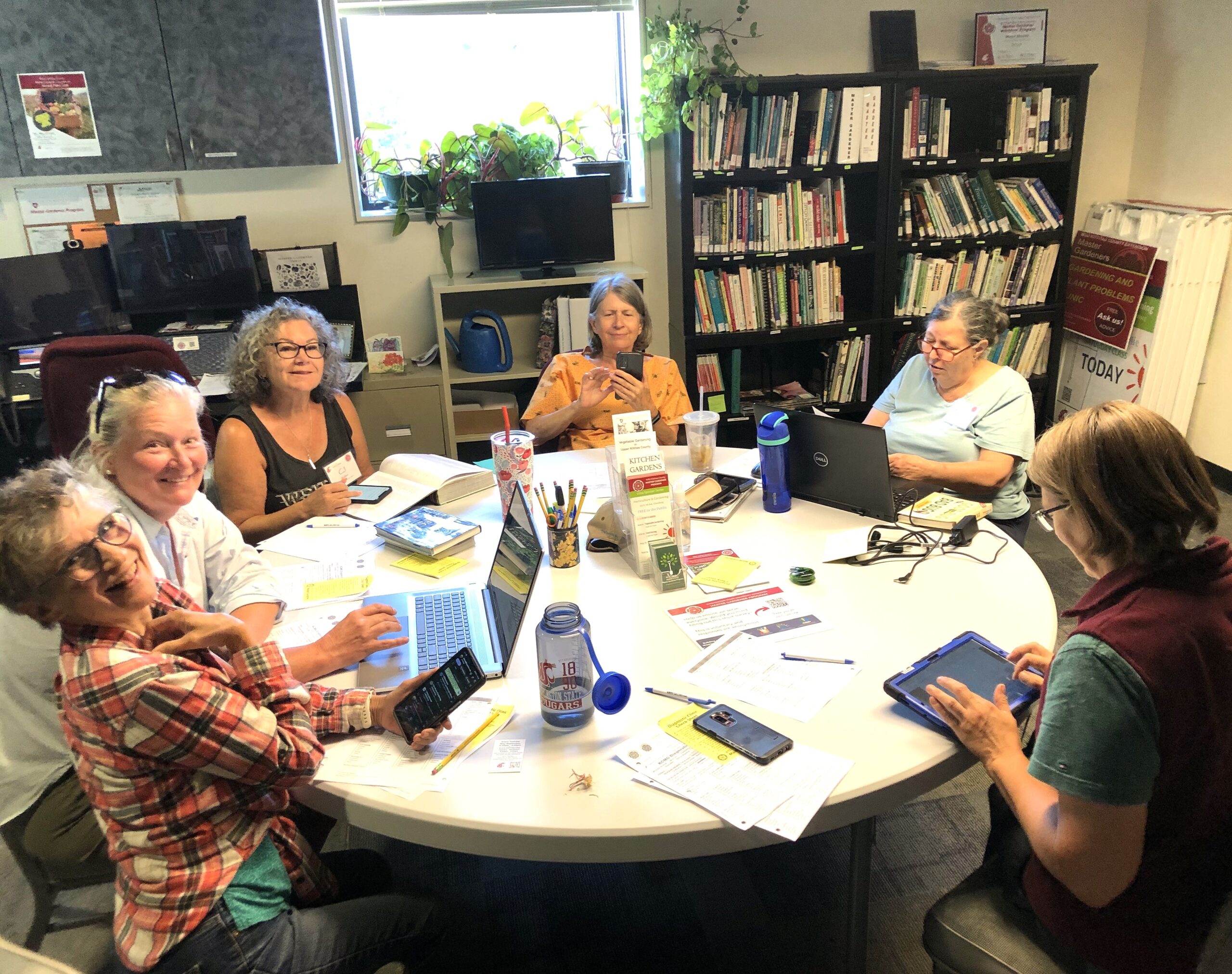 Six people sit in an office around a round table with their notes and devices.  Behind them there are bookshelves, educational materials, and plants in the window.