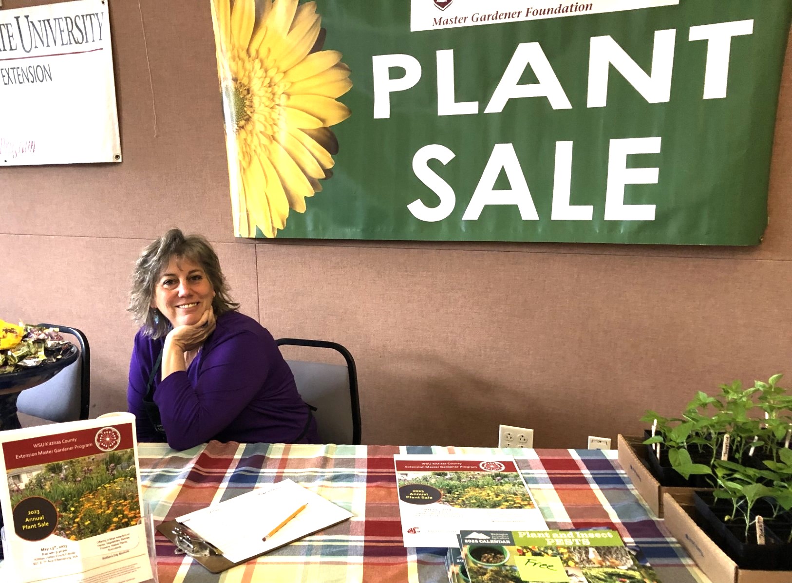 A Master Gardener volunteer sits behind a table at the annual plant sale.  Behind her is a large sign reading Plant Sale with a yellow flower at left.