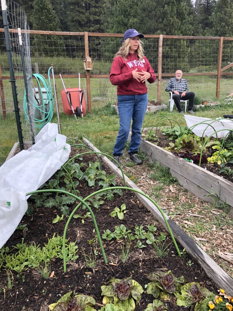 A Master Gardener wearing a baseball cap and hooded sweatshirt talks to others off-camera while standing in between raised garden beds.