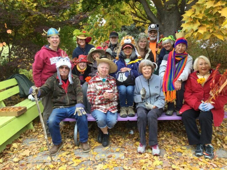 A group of master gardeners sit on a purple bench in under a tree with leaves turning yellow in the fall.  They are all wearing or carrying silly accessories, such as viking hats, animal hats, flowers, and bright scarves.