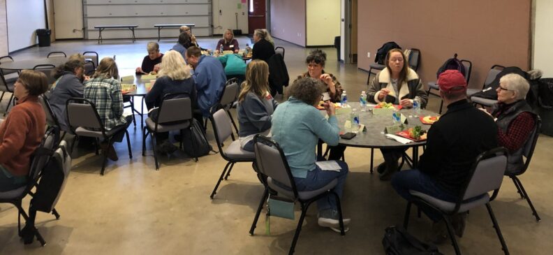 A group of Master Gardener trainees sit at round tables in an open teaching space.  They have a variety of items on the tables, including name tents, their lunches, water bottles, and other personal items.