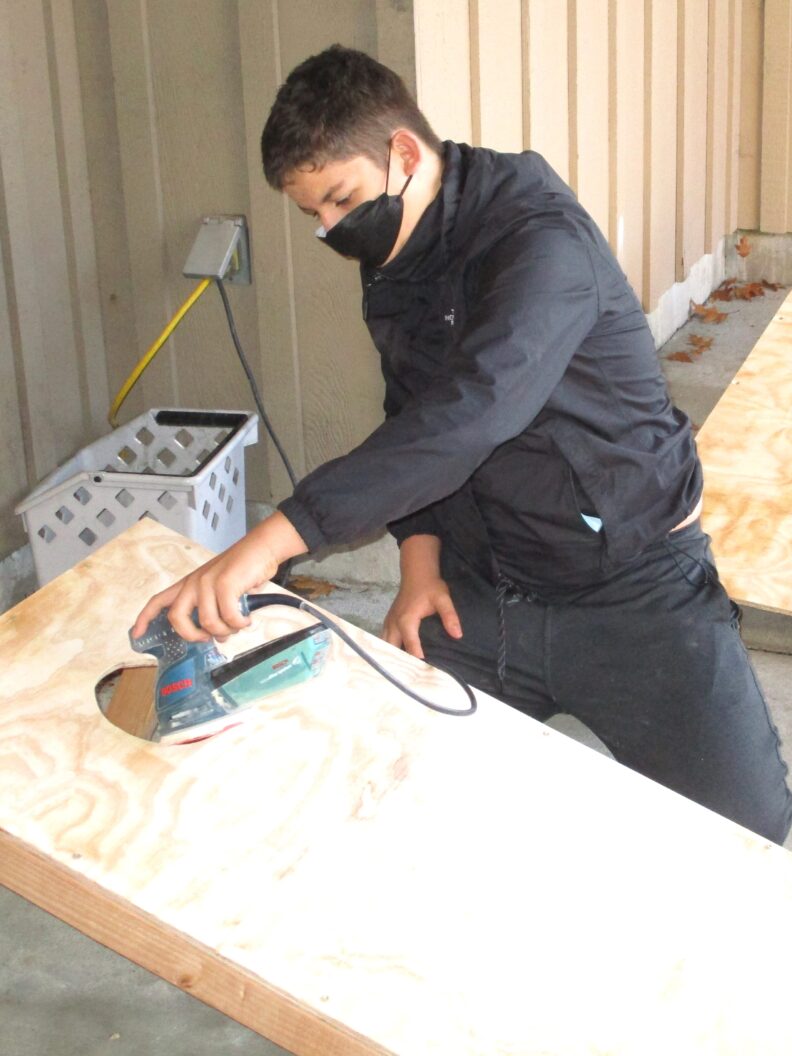 A youth member sanding while building a cornhole game set during a workshop