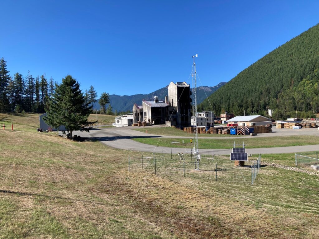 Weather instrumentation on a tower with buildings and mountains in the background.