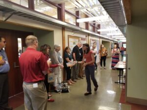 Woman speaks to group standing along wall in hallway.