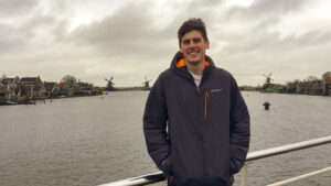 Young man stands at railing, river flanked by windmills and houses in background.