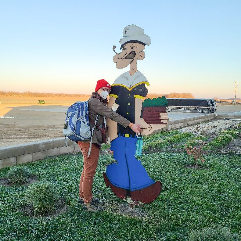 Woman wearing backpack hugs large plywood Popeye figure.