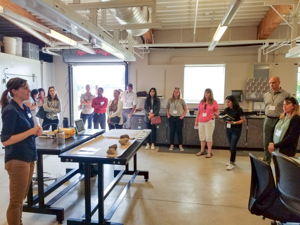 Woman speaks with group in large lab space; roll up door open to outside.