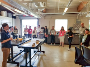 Woman speaks with group in large lab space; roll up door open to outside.