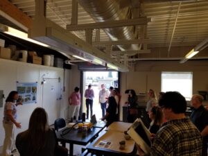 Woman speaks with group in large lab space; roll up door open to outside.