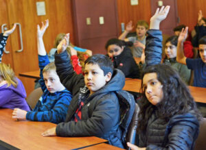 Students sit at desks with their hands raised.