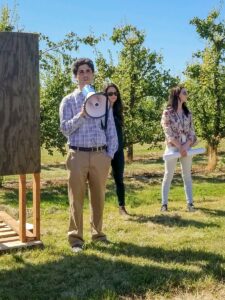 Travis Alexander presenting with a megaphone next to an orchard.