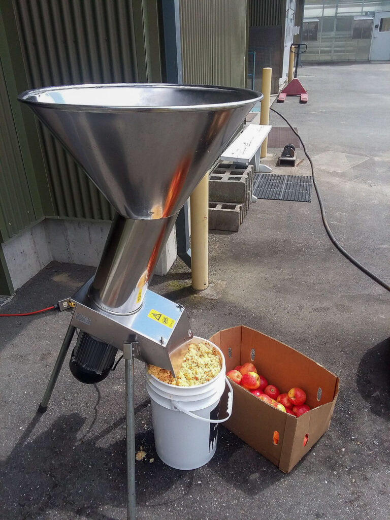 A box of apples sits next to a large stainless fruit grinder.