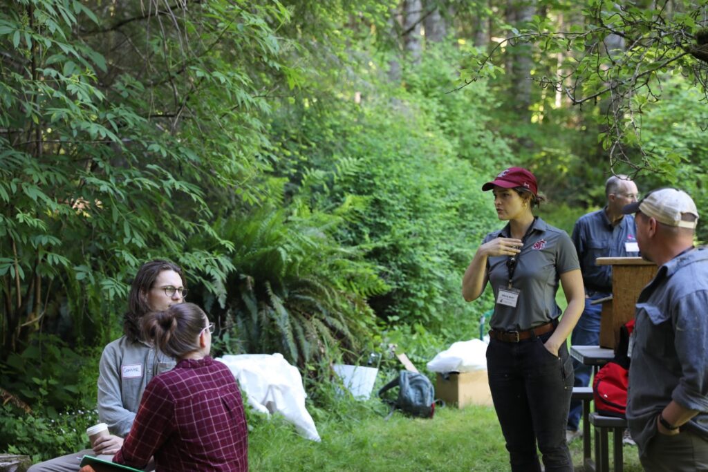 A woman talking with a dispersed group of people in a forest setting.