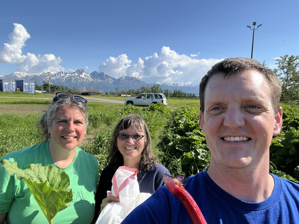 Three people in a field with large mountains in the distance. A man and a woman are holding stalks of rhubarb.
