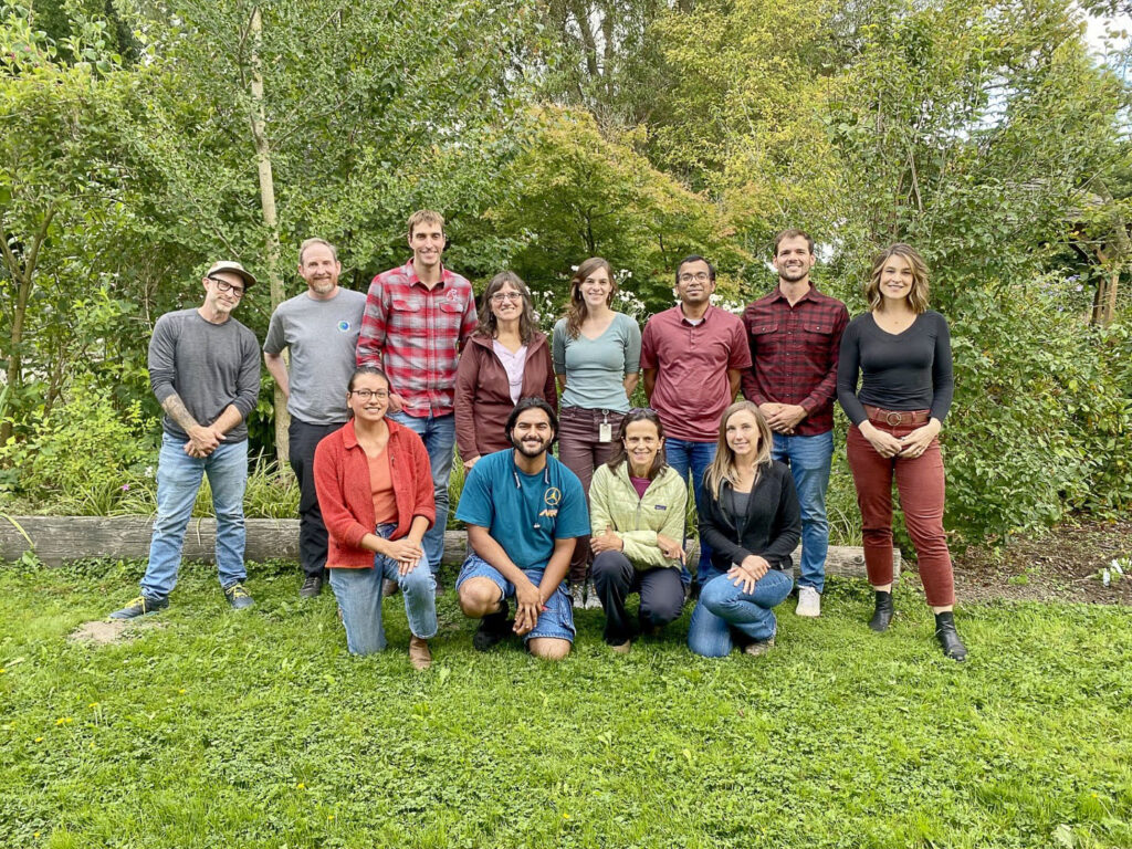 A group of 12 smiling people standing in front of a verdant garden.