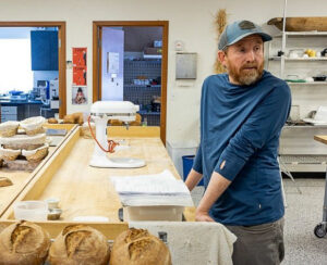 A man standing in a bakery kitchen next to a floured workbench, with loaves of bread and a large mixer.