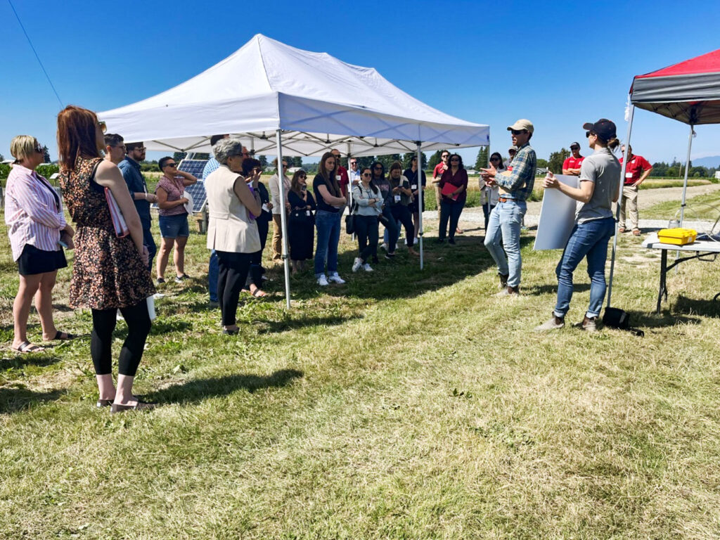 A large group of people stand clusters around a pop up tent listening to a researcher present findings in a sunny field under a pop up tent.