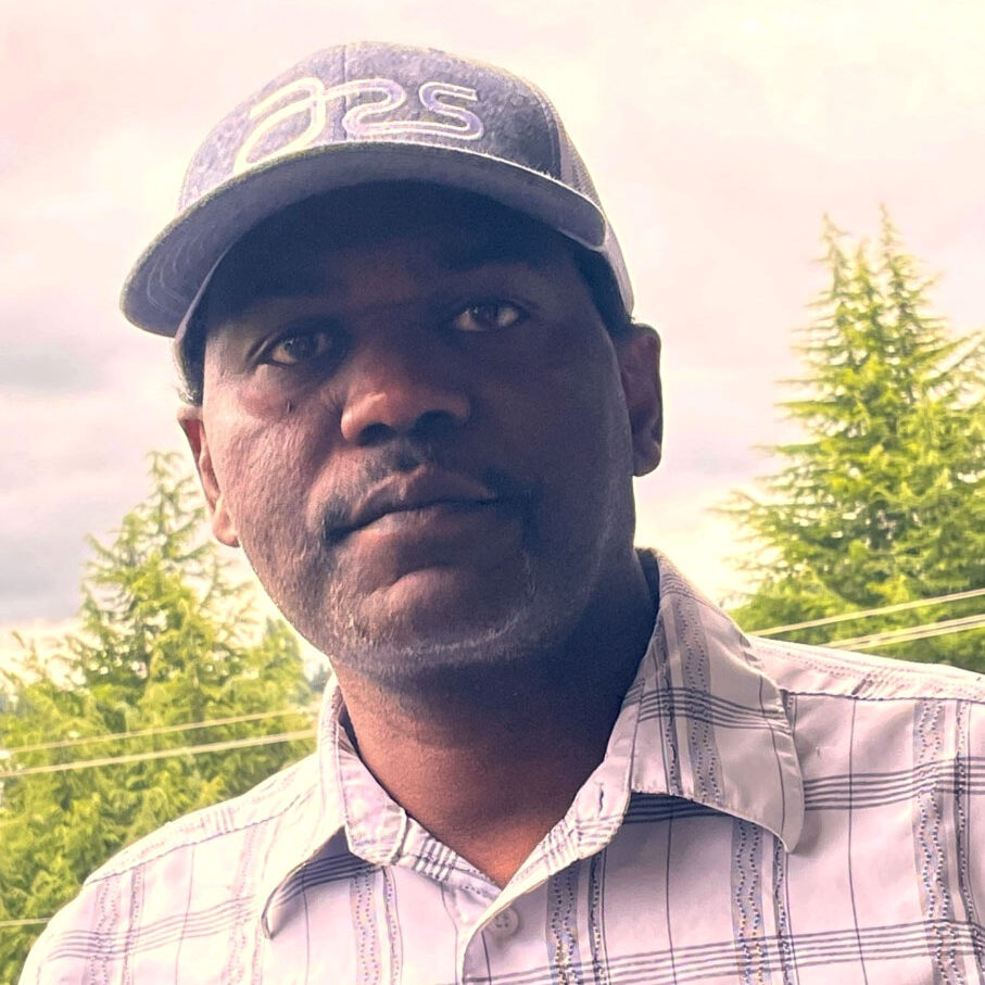 A photo of a man wearing a hat in front of a forest, looking at the camera.