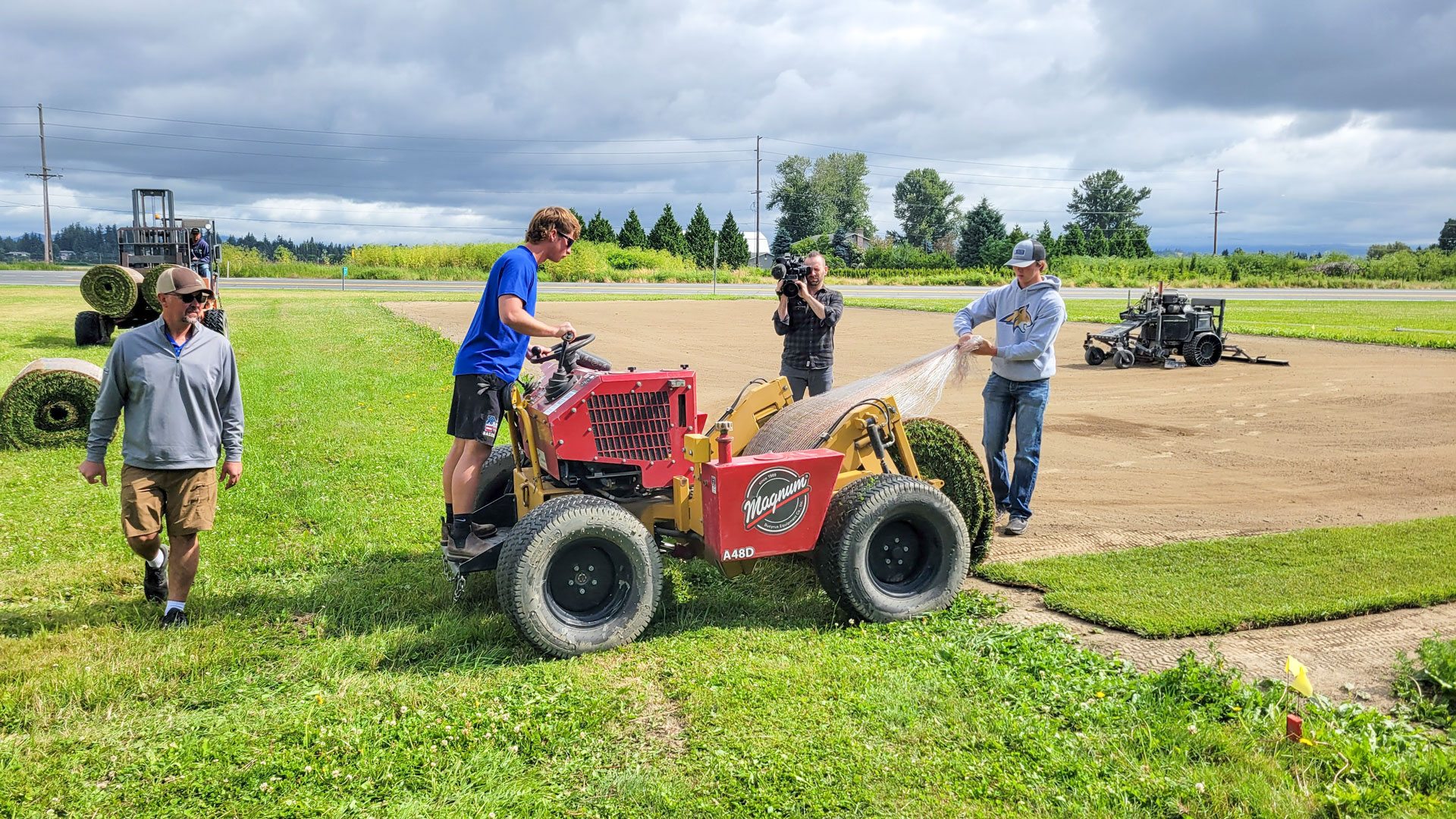 A cameraman films two young men unrolling sod using a machine.