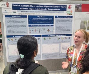 A woman in a bright floral shirt gestures in front of a large research poster on "Relative susceptability of northern highbush blueberry flower and fruit states to infection in Botrytis cinerea".
