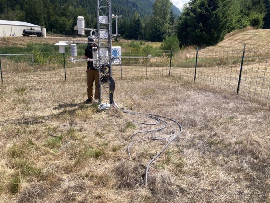 A person standing next to a metal tower with instruments and cords in a field of grass.