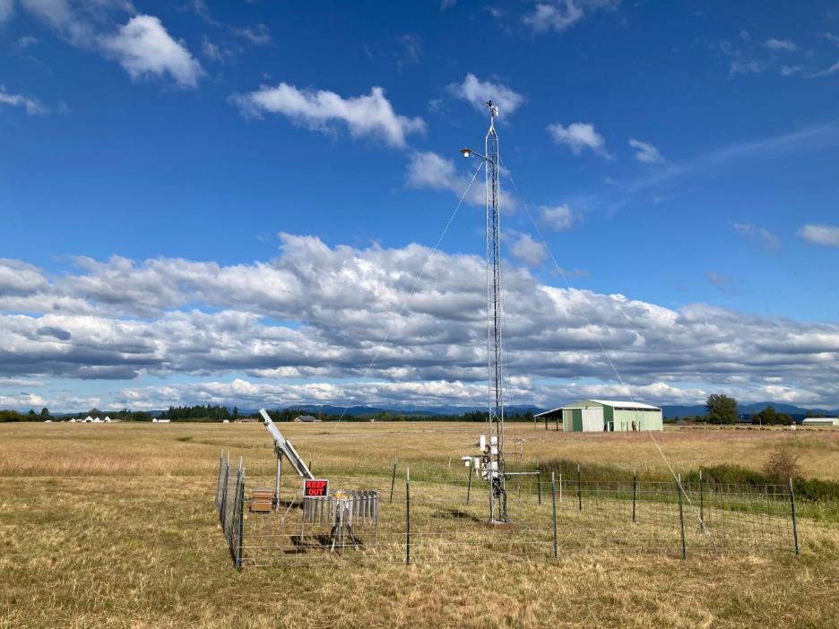 A metal tower with instruments surrounded by fencing on a wide open grassy field