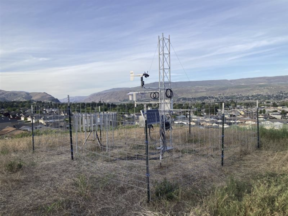 A wire fence around a metal tower with instruments and a solar panel with the city of Wenatchee in the background.