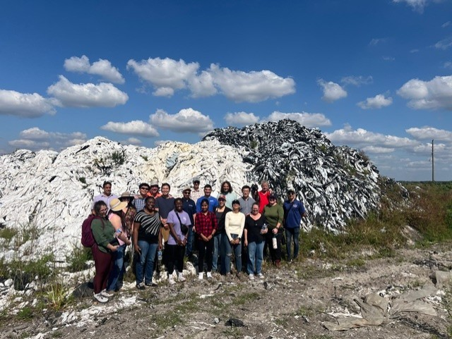 A group of 21 scientific researchers stand in front of a large pile of folded plastic mulch  sheets in white and black plastic.