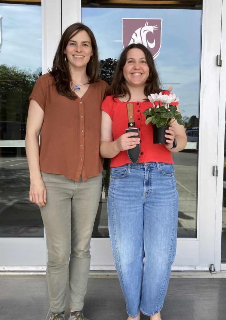 Two women standing in front of a doorway; the woman on the right is holding a spade and a potted plant.