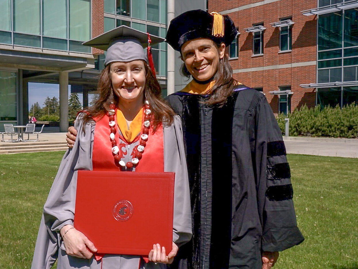 A woman in a graduation gown holding a red diploma and a woman in faculty regalia smiling at the camera.