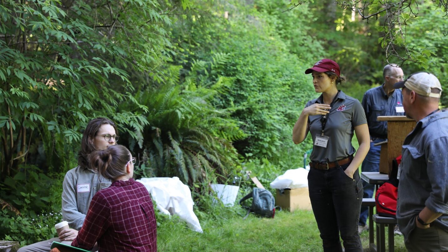 A woman talking with a dispersed group of people in a forest setting.