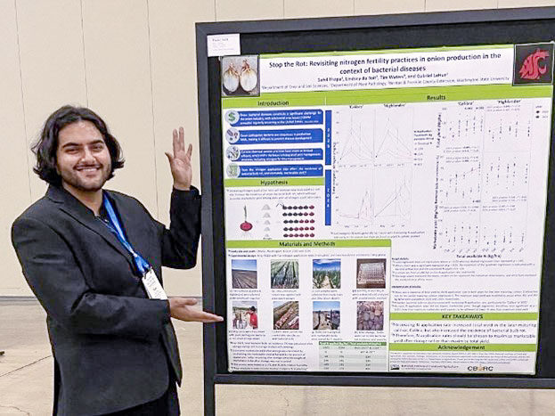 A man in a suit jacket motioning to a poster displayed next to him in a conference room.