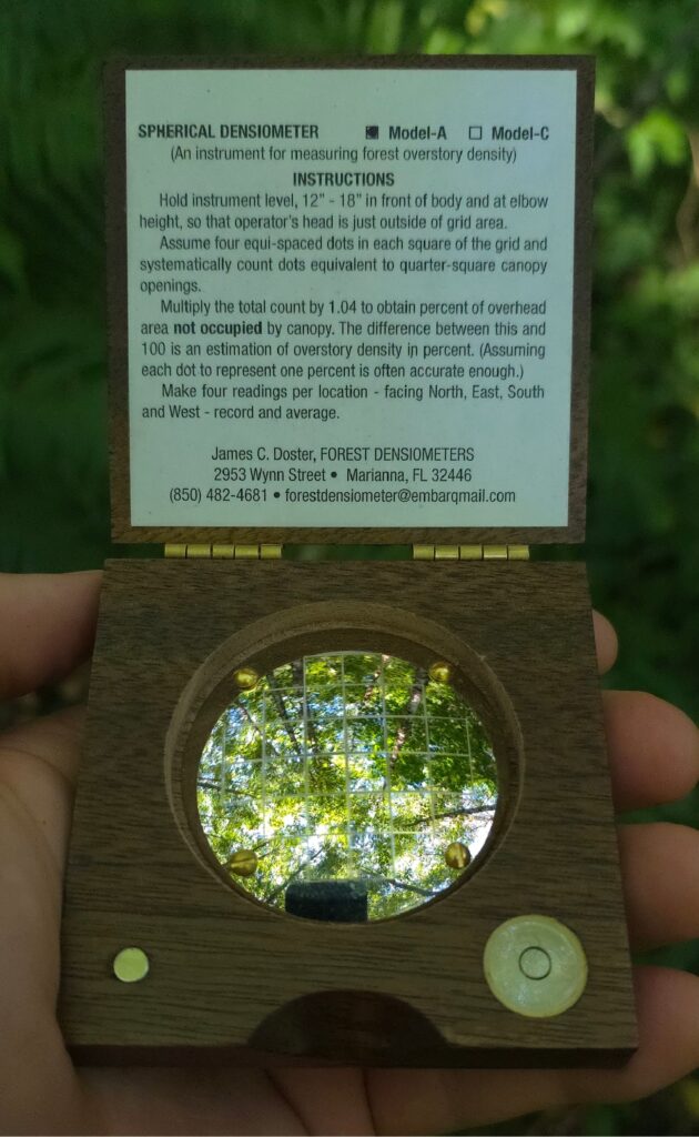A densiometer being held and used to determine shade levels in a forest. The densiometer shows a closed hardwood forest canopy with dapple sunlight coming through.