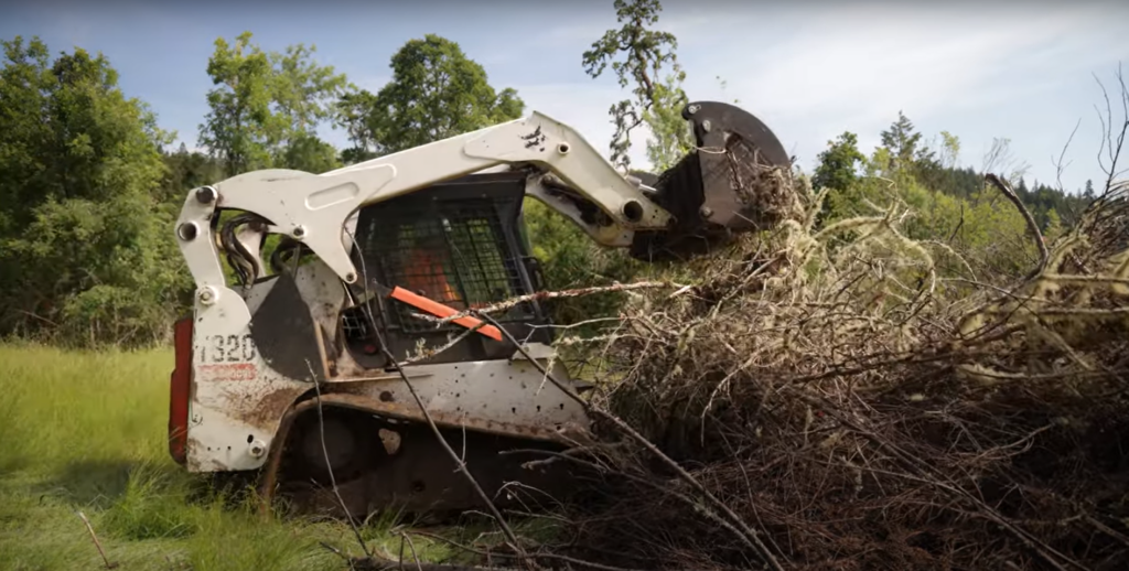 A skid steer, a large piece of machinery on tracks used to clear vegetation, is mulching woody debris.