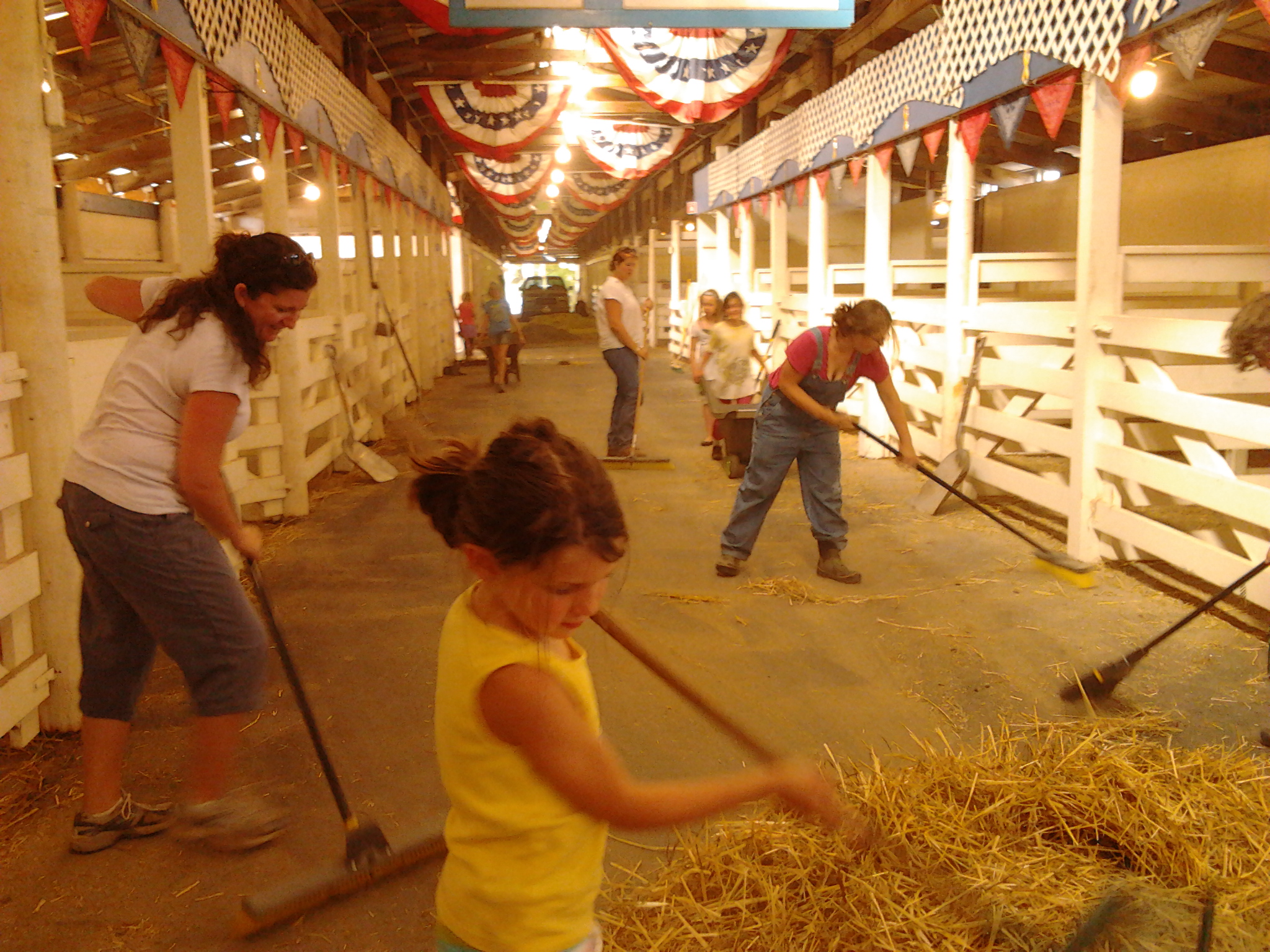 Many youth and adults sweeping hay in a barn. 