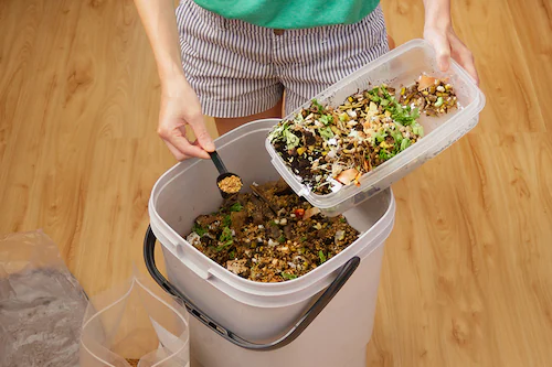 person with small scoop of bokashi bran being added to bokashi composting bucket.