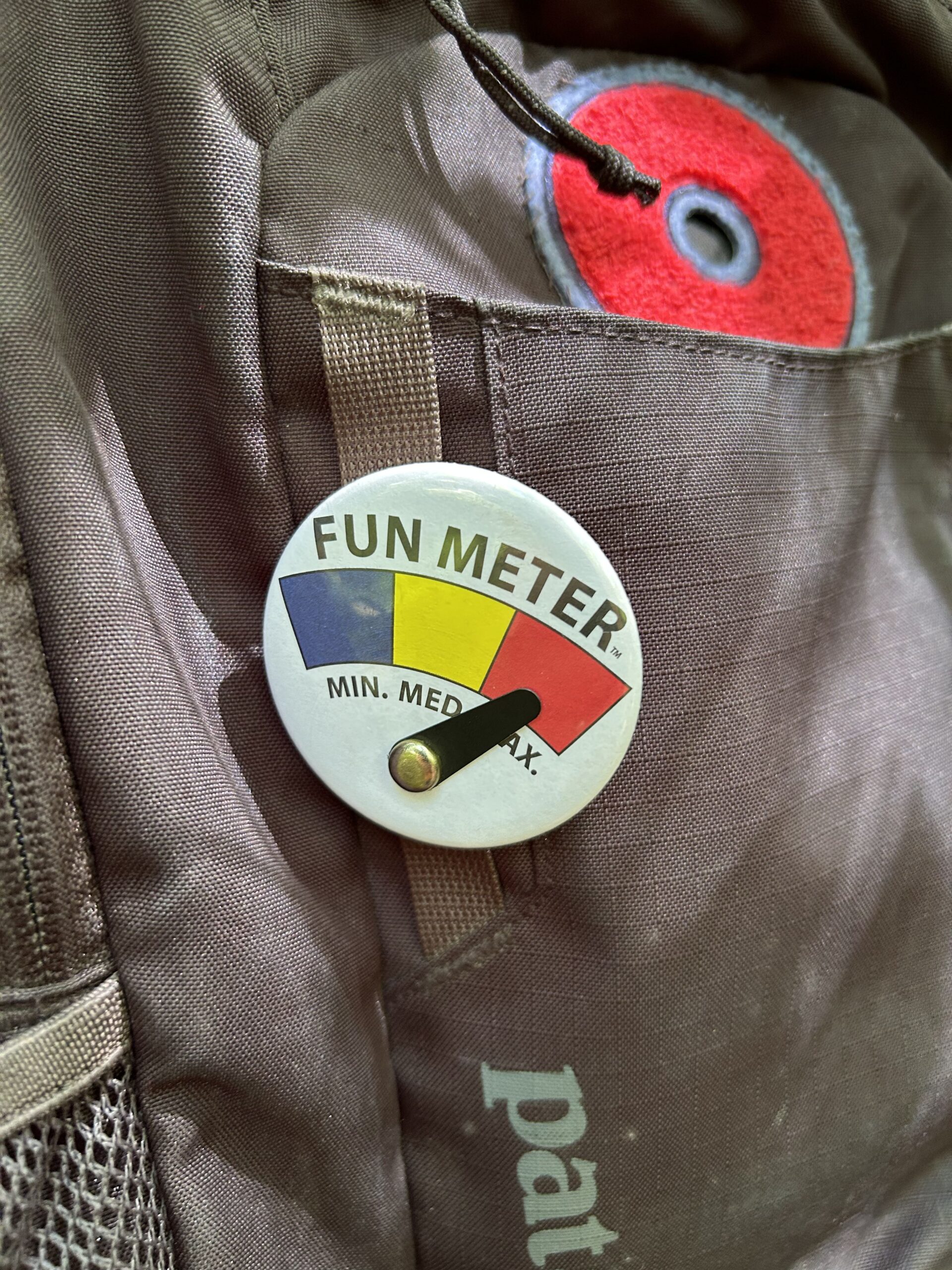Button that reads Fun Meter and has Min, Med., and Max. Pointer is pointed at "Max" in red. Button is on a backpack.